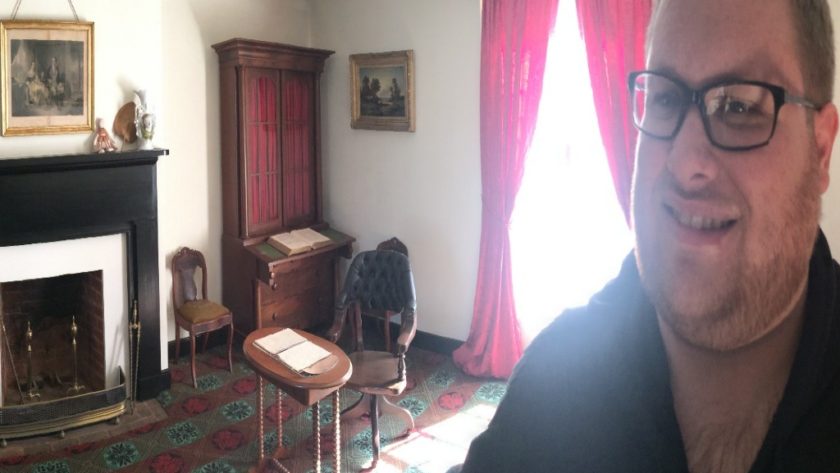 Douglas wearing a black sweatshirt standing in front and to the right of the chair where Ulysses S. Grant sat during the Surrender at Appomattox. There are red drapes behind the chair with sunlight coming through.